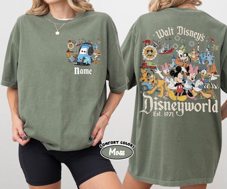 May include: Olive green t-shirt featuring a cartoon Mater design with the text "Name" on the front. The back of the shirt displays a colorful Disney World graphic with various characters and the text "Walt Disney's Disneyworld Est. 1971".