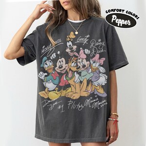 May include: Dark gray Comfort Colors t-shirt featuring a graphic of Disney characters including Mickey Mouse, Donald Duck, Goofy, Pluto, Minnie Mouse, and Daisy Duck. The shirt has the text "Comfort Colors Pepper" and character signatures.