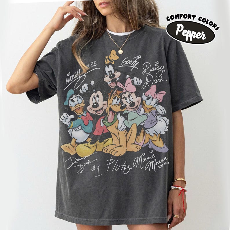 May include: Dark gray Comfort Colors t-shirt featuring a graphic of Disney characters including Mickey Mouse, Donald Duck, Goofy, Pluto, Minnie Mouse, and Daisy Duck. The shirt has the text "Comfort Colors Pepper" and character signatures.