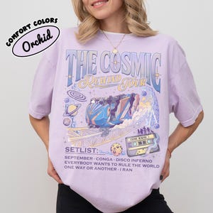 K&ouml;nnte beinhalten: Ein helllila T-Shirt mit einem grafischen Design mit dem Text "THE COSMIC REWIND TOUR". Das Design umfasst eine Achterbahn, Planeten und eine Kassette. Das Shirt listet auch eine Setlist auf.