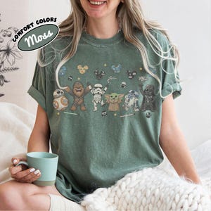 K&ouml;nnte beinhalten: Ein moosgr&uuml;nes T-Shirt mit einem Grafikdruck von Star Wars-Figuren, darunter Chewbacca, ein Sturmtruppler, Grogu, R2-D2, Darth Vader und BB-8, sowie kleine Symbole von Mickey-Maus-Ohren, Sternen und Lichtschwertern. Ein Etikett zeigt Comfort Colors Moss.