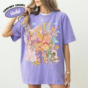 May include: A violet short-sleeve t-shirt featuring a graphic print of various animated characters in a collage style. The design includes the text "TS" at the top and a yellow sheriff star. The shirt has a crew neckline and a relaxed fit.