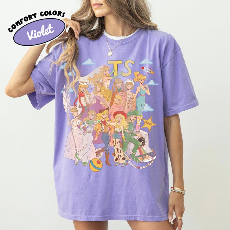 May include: A violet short-sleeve t-shirt featuring a graphic print of various animated characters in a collage style. The design includes the text "TS" at the top and a yellow sheriff star. The shirt has a crew neckline and a relaxed fit.