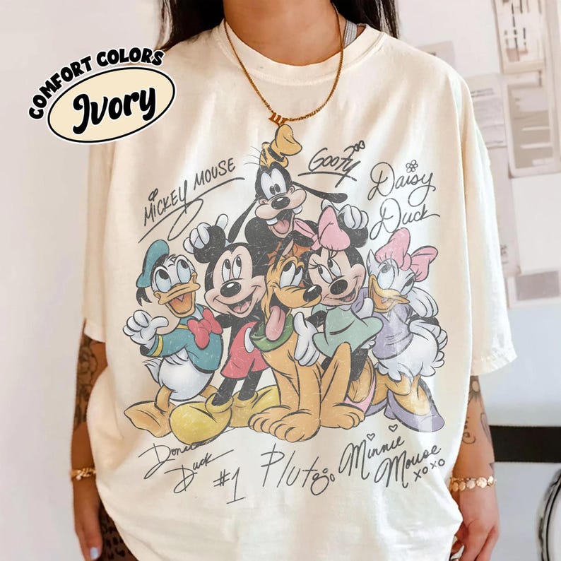 May include: Ivory-colored t-shirt featuring a graphic of Mickey Mouse, Minnie Mouse, Donald Duck, Goofy, Pluto, and Daisy Duck. The shirt has the words "Comfort Colors" and "Ivory" printed on it, along with character signatures.