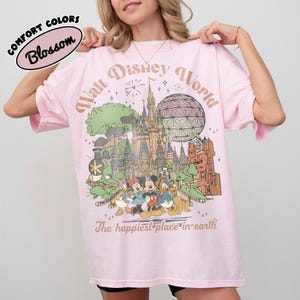 May include: A light pink t-shirt with a graphic of the Walt Disney World castle, characters, and the text "Walt Disney World" and "The happiest place on earth." The shirt also has the text "Comfort Colors Blossom."