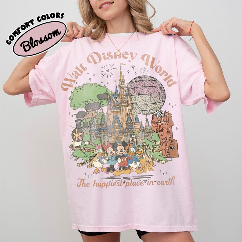 May include: A light pink t-shirt with a graphic of the Walt Disney World castle, characters, and the text "Walt Disney World" and "The happiest place on earth." The shirt also has the text "Comfort Colors Blossom."