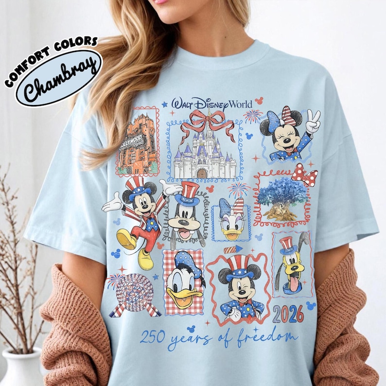 May include: Light blue t-shirt with a graphic featuring Disney characters and landmarks. The design includes Mickey Mouse, Minnie Mouse, Donald Duck, and Pluto in patriotic attire, along with the text "250 years of freedom" and "2026". The shirt has the text "Comfort Colors Chambray".