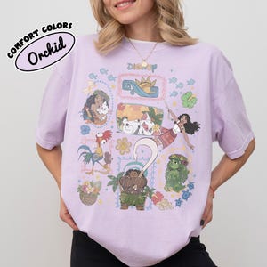 May include: Orchid-coloured t-shirt with a Disney graphic featuring characters from the movie Moana. The design includes illustrations of Moana, Maui, Hei Hei, Pua, and other elements from the film. The shirt is a Comfort Colors brand.