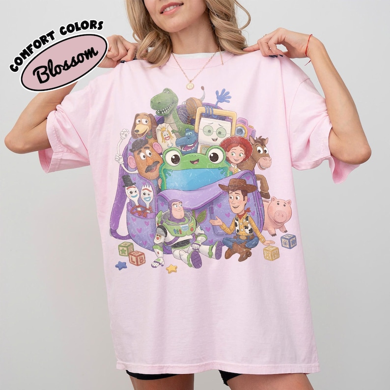 Pode incluir: Uma t-shirt rosa claro com uma impress&atilde;o gr&aacute;fica de v&aacute;rias personagens de brinquedos animados reunidas &agrave; volta de uma mochila roxa. O design inclui um sapo verde, um ranger espacial, um cowboy e outras figuras, com o texto Comfort Colors Blossom.