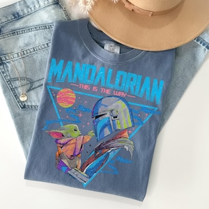 Może przedstawiać: Niebieska koszulka z grafiką The Mandalorian i Grogu, z napisem "MANDALORIAN - THIS IS THE WAY." Koszulka jest zestawiona z jasnymi dżinsami, brązowym kapeluszem i złotym naszyjnikiem.