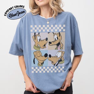 May include: Blue t-shirt with a graphic of Pluto in four panels, set against a checkered background. The shirt has the text "COMFORT COLORS Blue Jean" in a circular design.