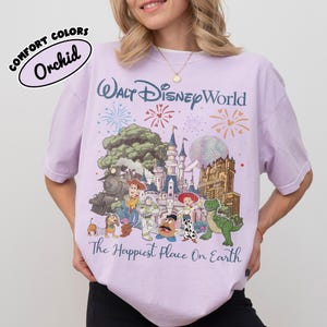 May include: Orchid Comfort Colors t-shirt featuring a Walt Disney World graphic. The design includes a castle, fireworks, and various Disney characters. The text "The Happiest Place On Earth" is printed below the graphic.