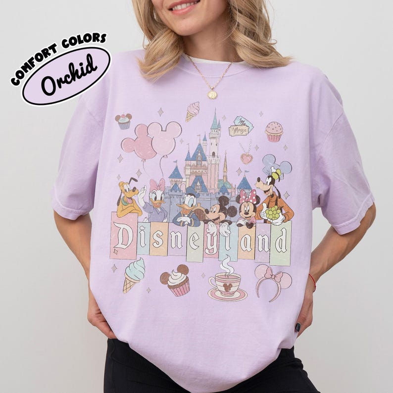 May include: A light purple t-shirt featuring a graphic print of Disneyland with Mickey Mouse, Minnie Mouse, Donald Duck, Daisy Duck, Goofy, and Pluto. The design includes a castle, balloons, cupcakes, ice cream, and the text Disneyland.