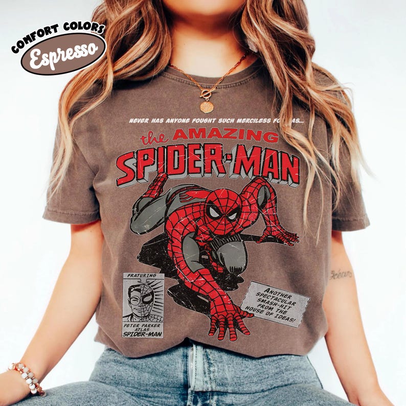 May include: An espresso-colored t-shirt featuring a vintage-style graphic of Spider-Man in a crouched pose. The text reads: Never has anyone fought such merciless foes, The Amazing Spider-Man, Featuring Peter Parker, and Another spectacular smash-hit.