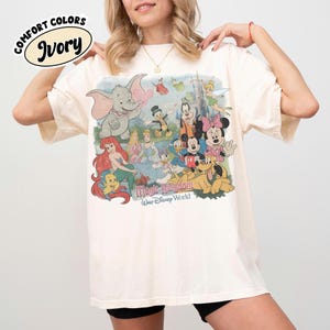 May include: Ivory-colored t-shirt featuring a colorful graphic of Disney characters, including Dumbo, Ariel, Mickey Mouse, and Minnie Mouse. The graphic is set against a backdrop of a castle and the words "Magic Kingdom" and "Walt Disney World".