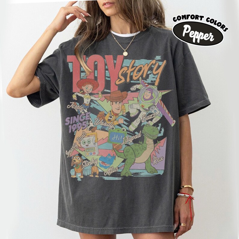 May include: A charcoal gray short-sleeve t-shirt featuring a vintage-style graphic print of Toy Story characters including Woody, Buzz Lightyear, Jessie, Rex, Slinky, and others. The text reads Toy Story, Since 1995, and Comfort Colors Pepper.
