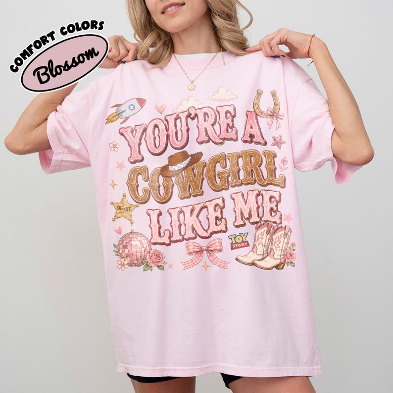 Puede incluir: Una camiseta rosa claro con el texto YOU'RE A COWGIRL LIKE ME en letras marrones y rosas. El gr&aacute;fico incluye un cohete, un sombrero de vaquero, una estrella de sheriff, una bola de discoteca, una herradura, un lazo y botas con el logo de Toy Story.