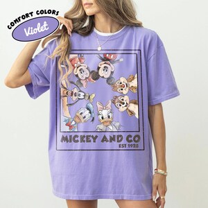 Puede incluir: Camiseta Violet Comfort Colors con un gr&aacute;fico de Mickey Mouse, Minnie Mouse, Donald Duck, Daisy Duck, Goofy y Chip y Chop. La camiseta tiene el texto "Mickey and Co. Est. 1928" en un rect&aacute;ngulo negro.
