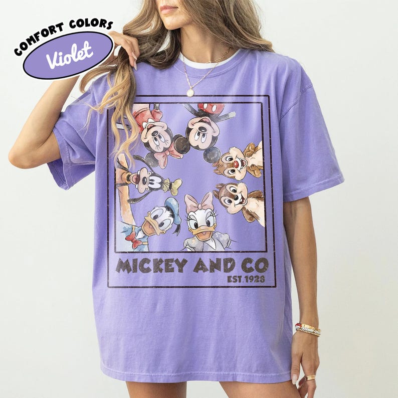 Puede incluir: Camiseta Violet Comfort Colors con un gr&aacute;fico de Mickey Mouse, Minnie Mouse, Donald Duck, Daisy Duck, Goofy y Chip y Chop. La camiseta tiene el texto "Mickey and Co. Est. 1928" en un rect&aacute;ngulo negro.