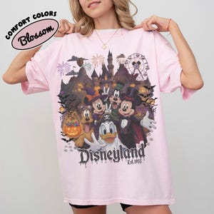 May include: A light pink t-shirt featuring a graphic print of Disney characters in Halloween costumes, including Mickey Mouse, Minnie Mouse, Donald Duck, Goofy, and Pluto. The design includes a castle, fireworks, and the text Disneyland Est. 1955.