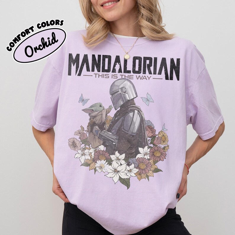 May include: Orchid-colored t-shirt featuring a graphic of The Mandalorian and Grogu surrounded by flowers and butterflies. The text "MANDALORIAN - THIS IS THE WAY" is printed above the image. The shirt has a Comfort Colors label.