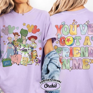 May include: A light purple t-shirt featuring a graphic of Toy Story characters including Woody, Buzz Lightyear, Jessie, Rex, and Hamm on the front. The back displays the colourful text "YOU'VE GOT A FRIEND IN ME" with a star icon and small hearts.