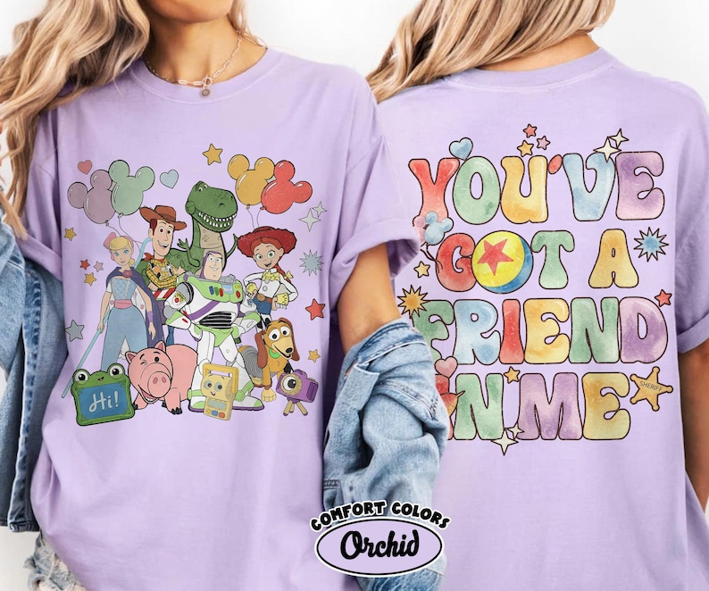 May include: A light purple t-shirt featuring a graphic of Toy Story characters including Woody, Buzz Lightyear, Jessie, Rex, and Hamm on the front. The back displays the colourful text "YOU'VE GOT A FRIEND IN ME" with a star icon and small hearts.