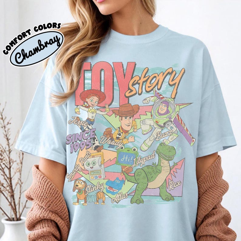 May include: A light blue t-shirt featuring a graphic print of Toy Story characters including Woody, Buzz Lightyear, Jessie, Rex, Slinky, and Smarty Pants. The design includes the text Toy Story, Since 1995, and character names in a colorful, retro-inspired layout.