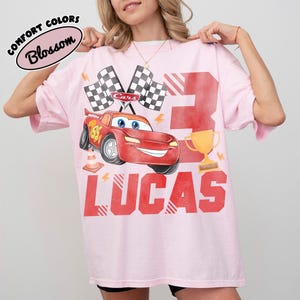 Puede incluir: Camiseta rosa con tem&aacute;tica de Cars. Presenta un coche de carreras rojo, banderas a cuadros, el n&uacute;mero 3, un trofeo dorado y el nombre "LUCAS" en rojo. La camiseta tambi&eacute;n tiene el texto "COMFORT COLORS Blossom".