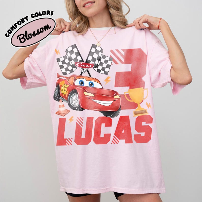 Puede incluir: Camiseta rosa con tem&aacute;tica de Cars. Presenta un coche de carreras rojo, banderas a cuadros, el n&uacute;mero 3, un trofeo dorado y el nombre "LUCAS" en rojo. La camiseta tambi&eacute;n tiene el texto "COMFORT COLORS Blossom".