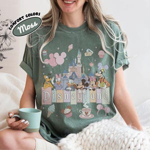 May include: A moss green t-shirt featuring a graphic print of a castle, Mickey Mouse, Minnie Mouse, Donald Duck, Daisy Duck, Goofy, and Pluto. The design includes pastel-colored balloons, cupcakes, ice cream, and the text Disneyland.