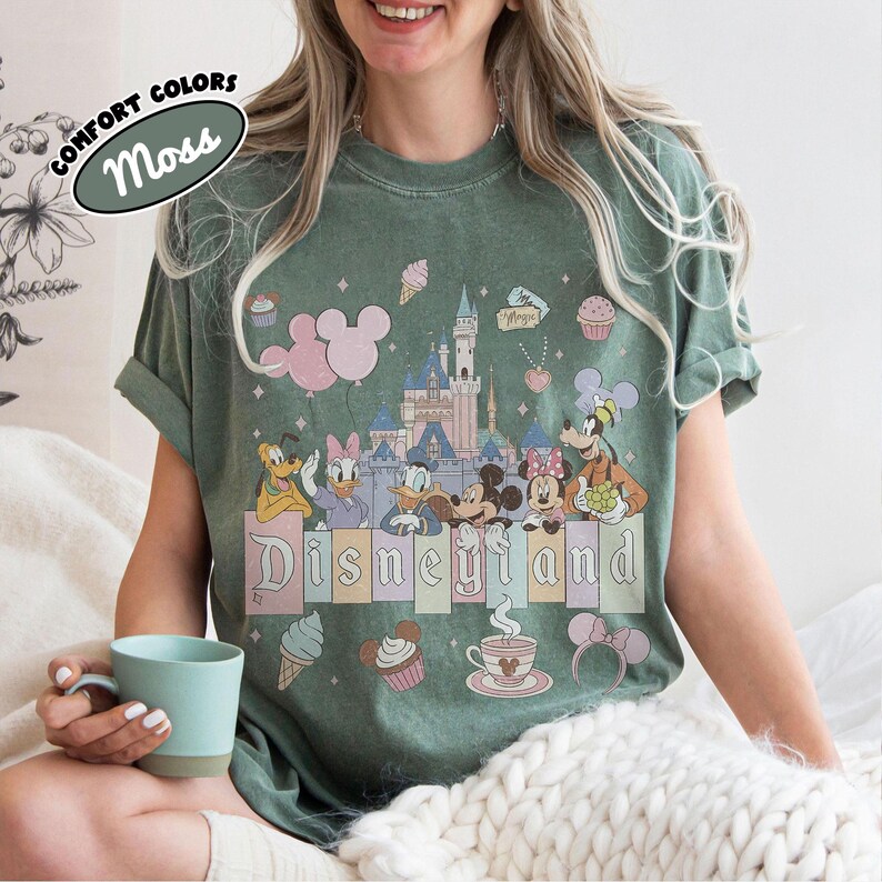 May include: A moss green t-shirt featuring a graphic print of a castle, Mickey Mouse, Minnie Mouse, Donald Duck, Daisy Duck, Goofy, and Pluto. The design includes pastel-colored balloons, cupcakes, ice cream, and the text Disneyland.