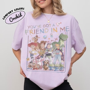 May include: A light purple short-sleeve t-shirt featuring a graphic print of various animated toy characters. The text "YOU'VE GOT A FRIEND IN ME" is printed above the characters in a colorful, distressed font. The shirt has a crew neckline.