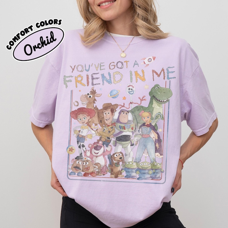 May include: A light purple short-sleeve t-shirt featuring a graphic print of various animated toy characters. The text "YOU'VE GOT A FRIEND IN ME" is printed above the characters in a colorful, distressed font. The shirt has a crew neckline.