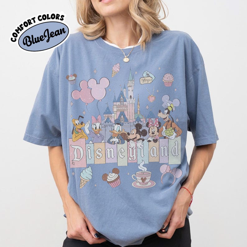 May include: A blue jean colored t-shirt featuring a graphic print of Disneyland with a castle, Mickey Mouse, Minnie Mouse, Donald Duck, Daisy Duck, Goofy, and Pluto. The design includes pastel-colored balloons, cupcakes, and a teacup.
