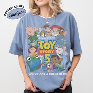 Op de afbeelding: Een blauw T-shirt met korte mouwen voorzien van een grafische print van diverse Toy Story-personages, de tekst Disney Pixar Toy Story 5 en de zin You've Got A Friend In Me. Een Comfort Colors Blue Jean-logo is linksboven zichtbaar.