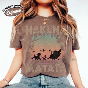 May include: Espresso-coloured t-shirt with the words "HAKUNA MATATA" in a distressed font. The design features silhouettes of Simba, Timon, Pumbaa, and a bird, set against a jungle backdrop. The shirt has a vintage, worn appearance.