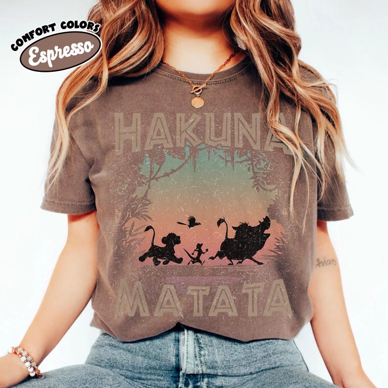 May include: Espresso-coloured t-shirt with the words "HAKUNA MATATA" in a distressed font. The design features silhouettes of Simba, Timon, Pumbaa, and a bird, set against a jungle backdrop. The shirt has a vintage, worn appearance.