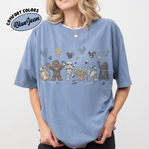K&ouml;nnte beinhalten: Ein blaues Kurzarm-T-Shirt mit einem Grafikdruck von Star Wars-Figuren wie Chewbacca, einem Stormtrooper, Grogu, R2-D2 und Darth Vader, die Luftballons in Form von Mickey-Mouse-Ohren halten. Der Text COMFORT COLORS BlueJean ist zu sehen.