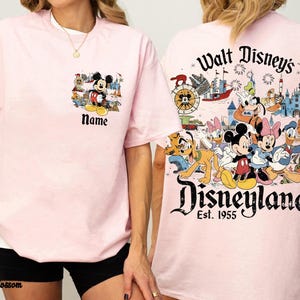 Peut inclure: T-shirt rose clair avec des motifs de Mickey Mouse et Disneyland. Le devant pr&eacute;sente un petit motif de Mickey Mouse avec le mot "Name" en dessous. Le dos affiche une grande sc&egrave;ne de Disneyland avec divers personnages Disney.