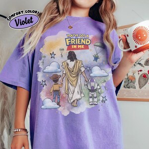 May include: Violet Comfort Colors t-shirt with a graphic featuring the text "YOU'VE GOT A FRIEND IN ME." The design includes illustrations of Jesus, Woody, Buzz Lightyear, and an alien, set against a colorful background with clouds and stars.