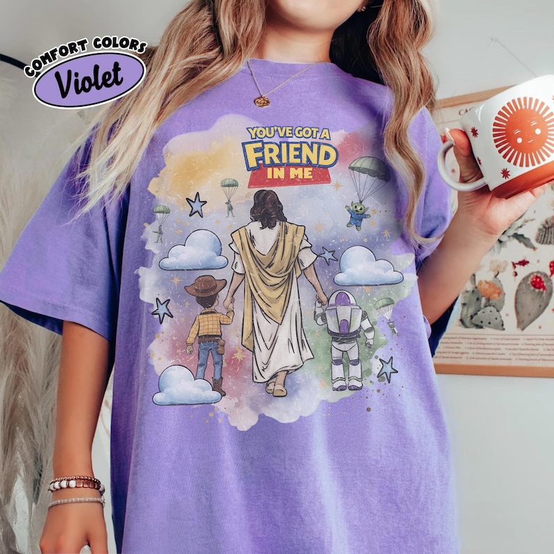 May include: Violet Comfort Colors t-shirt with a graphic featuring the text "YOU'VE GOT A FRIEND IN ME." The design includes illustrations of Jesus, Woody, Buzz Lightyear, and an alien, set against a colorful background with clouds and stars.