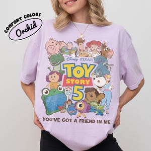 Op de afbeelding: Een lichtpaars t-shirt met een grafische print van Toy Story-personages, waaronder Woody, Buzz Lightyear en Jessie. De tekst luidt Disney Pixar Toy Story 5 en You've Got A Friend In Me. Een label in de hoek vermeldt Comfort Colors Orchid.