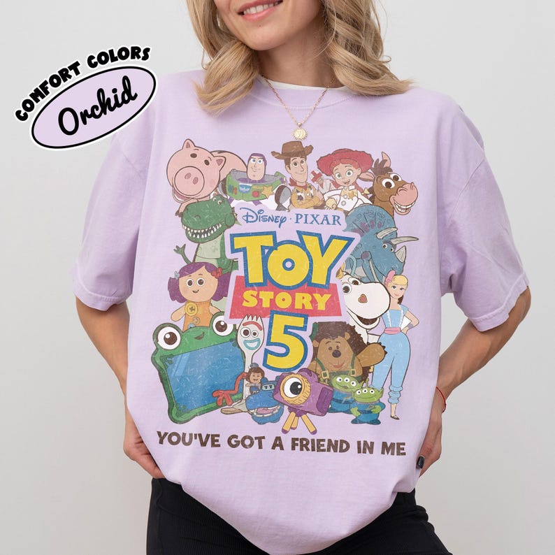 Op de afbeelding: Een lichtpaars t-shirt met een grafische print van Toy Story-personages, waaronder Woody, Buzz Lightyear en Jessie. De tekst luidt Disney Pixar Toy Story 5 en You've Got A Friend In Me. Een label in de hoek vermeldt Comfort Colors Orchid.