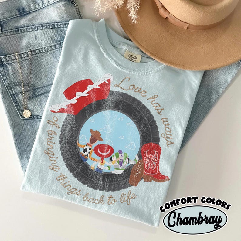 May include: A light blue t-shirt featuring a graphic of a black tire framing cartoon characters, a red cowboy hat, and a red cowboy boot. The text reads Love has ways of bringing things back to life. The shirt is placed near blue jeans and a tan hat.