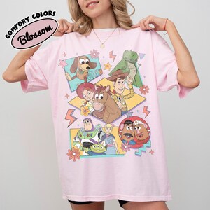 Puede incluir: Camiseta rosa claro con un gr&aacute;fico colorido de personajes de la pel&iacute;cula animada Toy Story. La camiseta tiene mangas cortas y el texto "COMFORT COLORS Blossom" en un &oacute;valo negro en la parte superior izquierda.