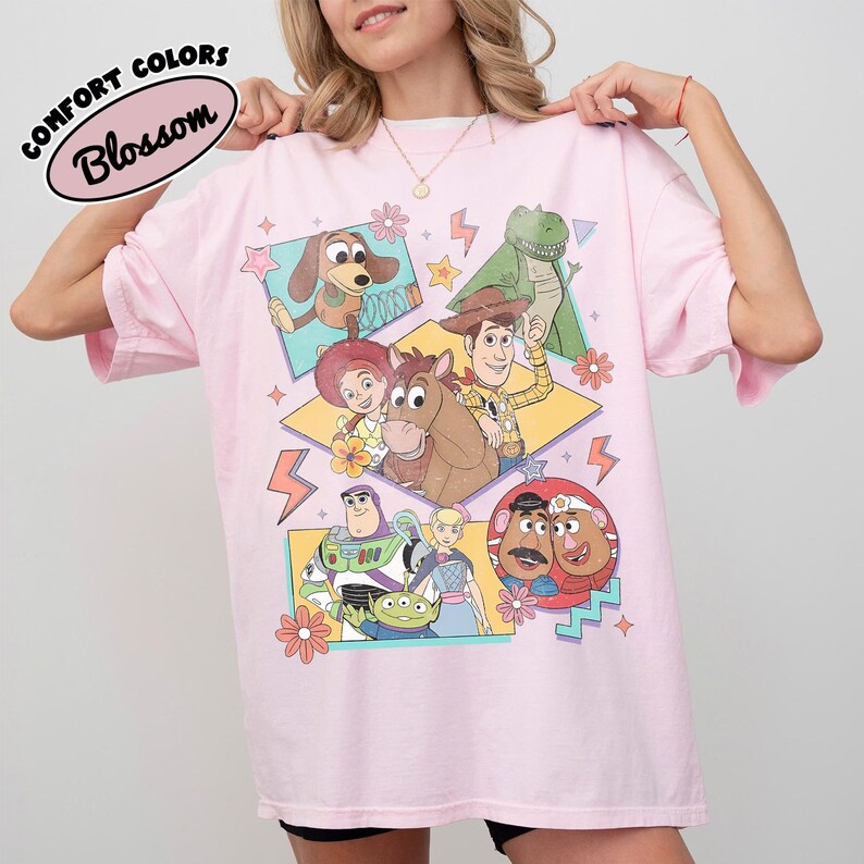 Puede incluir: Camiseta rosa claro con un gr&aacute;fico colorido de personajes de la pel&iacute;cula animada Toy Story. La camiseta tiene mangas cortas y el texto "COMFORT COLORS Blossom" en un &oacute;valo negro en la parte superior izquierda.