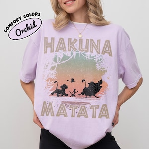 May include: A lilac t-shirt with the words "HAKUNA MATATA" in a tan font. The shirt features a graphic of characters from The Lion King, including Simba, Timon, Pumbaa, and Zazu. The shirt has a distressed appearance.