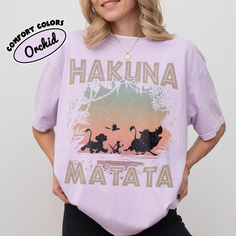 May include: A lilac t-shirt with the words "HAKUNA MATATA" in a tan font. The shirt features a graphic of characters from The Lion King, including Simba, Timon, Pumbaa, and Zazu. The shirt has a distressed appearance.