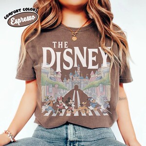 Może przedstawiać: Koszulka w kolorze espresso z napisem "THE DISNEY" nad kolorową ilustracją postaci Disneya przechodzących przez ulicę przed zamkiem. Koszulka ma wygląd vintage.